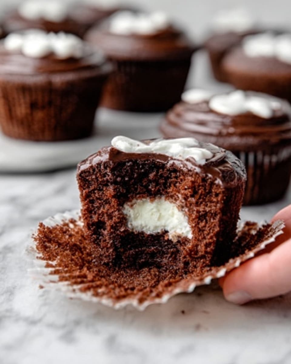 A close-up shot of a chocolate cupcake with a bite taken out of it, showing a white creamy filling inside. The cupcake has a moist, dark brown cake base and is topped with a smooth, dark chocolate frosting layer with a bit of white icing decoration on top. Several similar cupcakes, blurred in the background, sit on a white marbled surface. A woman's hand is gently holding the bitten cupcake from the side. photo taken with an iphone --ar 4:5 --v 7