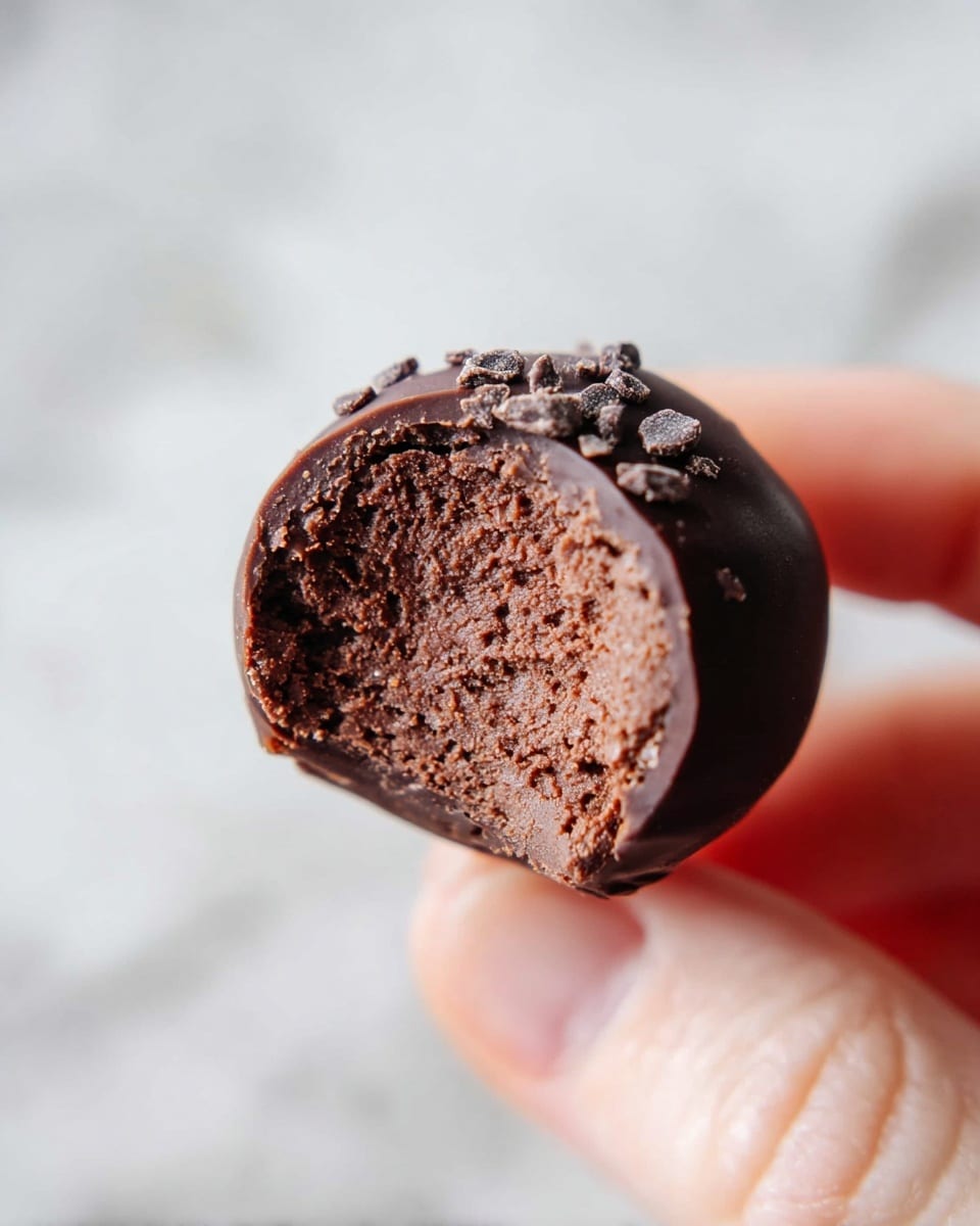 A close-up view shows a half-eaten round chocolate truffle held by a woman's hand between thumb and forefinger. The outer layer is a smooth and glossy dark brown chocolate shell sprinkled lightly with small chocolate crumbs. Inside, the truffle has a dense, slightly grainy texture with a richer and thicker chocolate filling in a lighter brown shade. The background is a soft white marbled texture that makes the chocolate stand out clearly. photo taken with an iphone --ar 4:5 --v 7