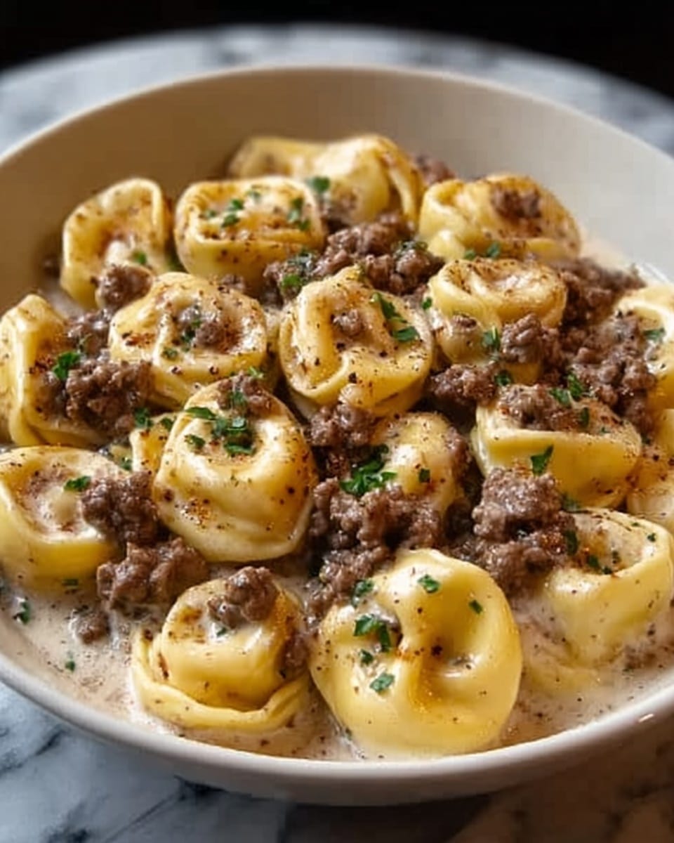 A close-up of a white bowl filled with creamy tortellini pasta topped with browned ground meat and finely chopped herbs. The tortellini are a pale yellow color with a smooth texture, arranged in a loose pile. The sauce underneath is creamy and light-colored, slightly visible between the pasta pieces. Small green herb bits are sprinkled evenly across the dish, adding a touch of freshness. The white marbled surface beneath the bowl contrasts softly with the darker colors of the meat and pasta. photo taken with an iphone --ar 4:5 --v 7