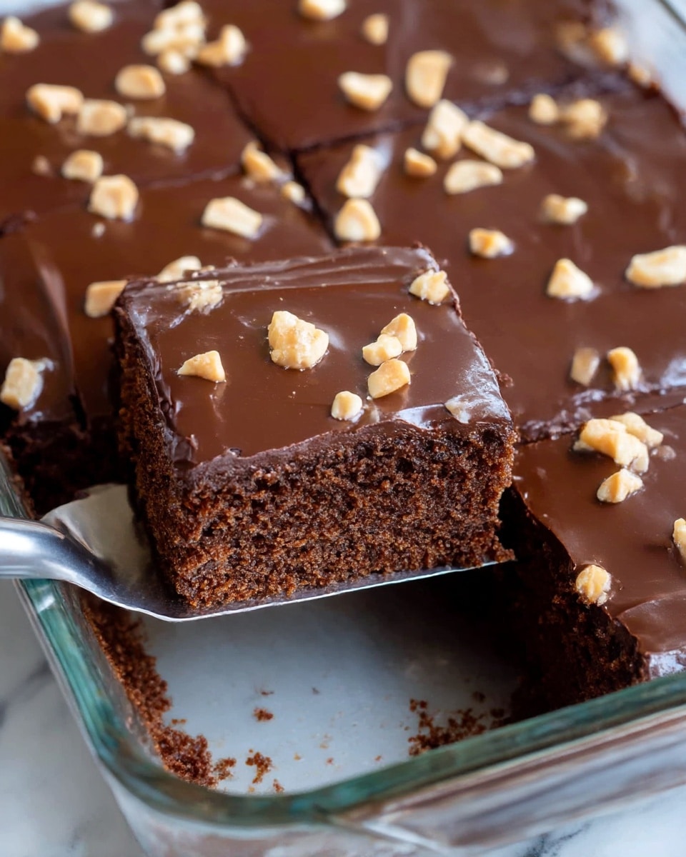 This image shows a chocolate sheet cake with two visible layers: the bottom layer is a thick, moist, dark brown cake with a soft crumb texture; the top layer is a shiny, smooth dark chocolate frosting covering the whole cake evenly. There are scattered small pieces of light tan chopped peanuts on the frosting for decoration. One slice has been cut and is being lifted by a metal spatula, showing the cake's interior. The cake is in a clear glass baking dish, placed on a surface with a white marbled texture. photo taken with an iphone --ar 4:5 --v 7