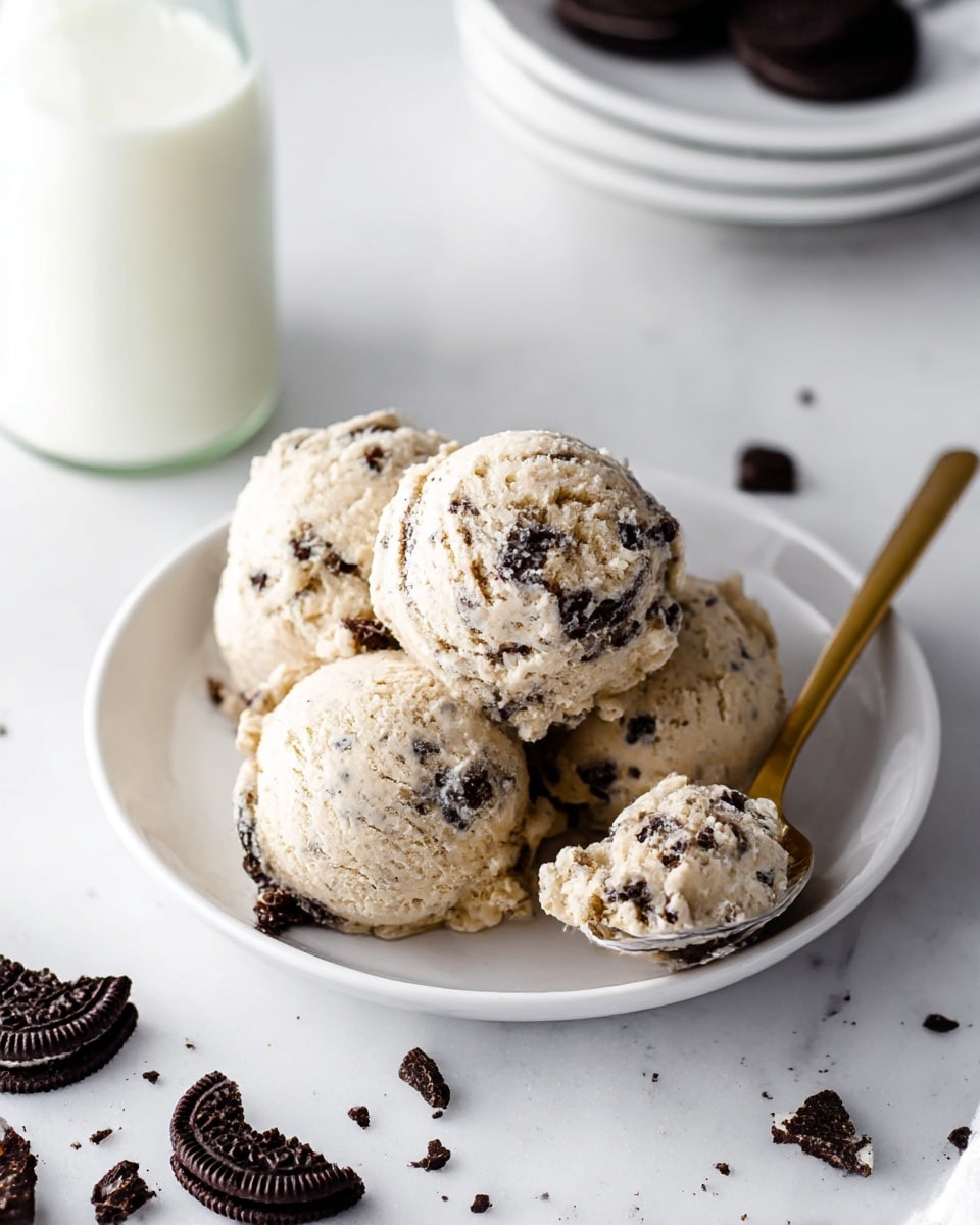 A white plate holds four scoops of cookies and cream ice cream, each scoop pale beige with dark chocolate cookie chunks mixed evenly throughout. The ice cream looks creamy with a textured surface showing cookie pieces scattered on top and inside. To the right of the plate, a golden spoon holds a small scoop of the ice cream. Around the plate on a white marbled surface, there are broken pieces of dark chocolate sandwich cookies with white cream filling visible. In the foreground, a blurred glass of milk is partially visible, and to the left, a small stack of white plates is slightly out of focus. photo taken with an iphone --ar 4:5 --v 7