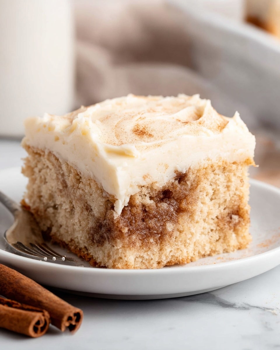 A square piece of cinnamon cake sits on a white plate, showing two clear layers: the bottom is a light brown, moist cake with swirls of darker cinnamon filling, giving it a textured look, while the top layer is thick, smooth, and creamy pale off-white frosting that evenly covers the cake. The plate rests on a white marbled texture, and a cinnamon stick is placed nearby, adding to the cozy feel of the scene. The background is softly blurred with light neutral tones, enhancing the cake's detailed textures. photo taken with an iphone --ar 4:5 --v 7