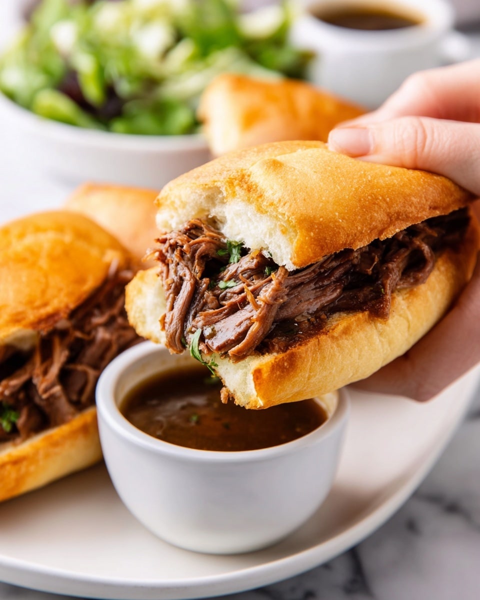 A close-up of a sandwich held by a woman's hand, showing a soft, golden-brown bun with a slightly crispy outside. Inside the bun, there is a thick layer of shredded dark brown beef piled high with visible juice and texture. The sandwich is being dipped into a small white cup of rich, dark brown broth placed on a white plate. In the background, there is another sandwich and a bowl of green leafy salad, both sitting on a white marbled surface. photo taken with an iphone --ar 4:5 --v 7