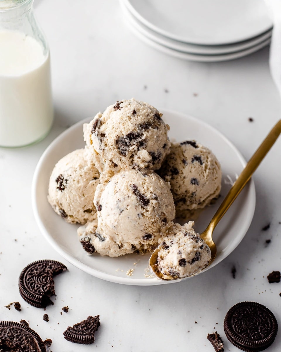 The image shows four scoops of light beige cream with dark chocolate cookie chunks mixed throughout, placed on a white plate. The scoops have a rough texture with visible cookie pieces embedded in all of them, sitting close together in the center of the plate. A gold spoon rests on the right side of the plate, holding a small scoop of the cream. Around the plate, there are broken pieces of dark chocolate sandwich cookies scattered on a white marbled surface. In the background to the left, part of a glass bottle filled with milk and a stack of white plates are visible, all set on the same white marbled surface. Photo taken with an iphone --ar 4:5 --v 7