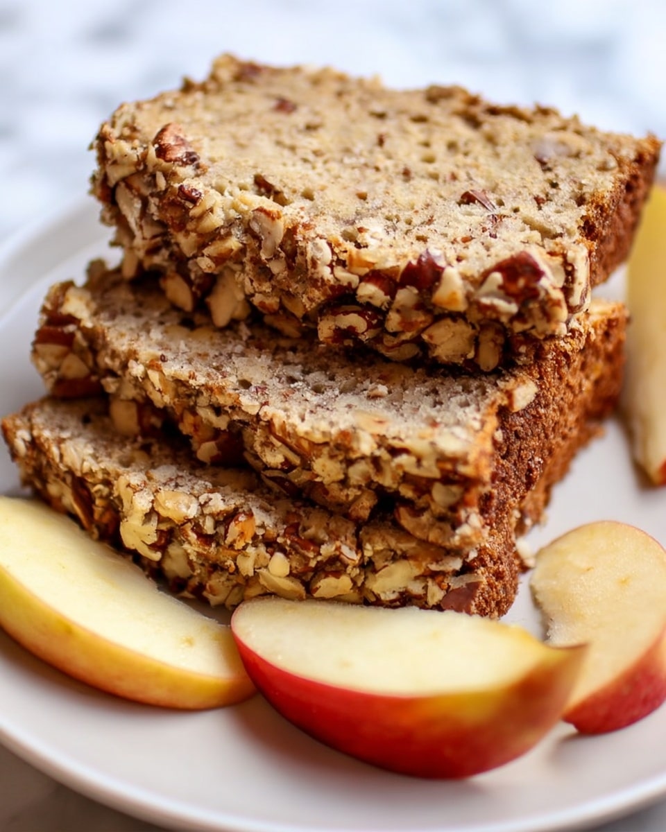 The image shows three slices of nutty bread stacked on a white plate, each slice thick and textured with visible chopped nuts embedded throughout. The top slice has a crunchy, golden-brown crust covered with more nuts, creating a rough, uneven surface, while the inside is soft, speckled with small holes and nut pieces. Around the bread slices, there are fresh apple wedges with red and yellow skin adding a fresh contrast. The plate rests on a white marbled surface that brightens the scene, giving a clean and fresh look. Photo taken with an iphone --ar 4:5 --v 7
