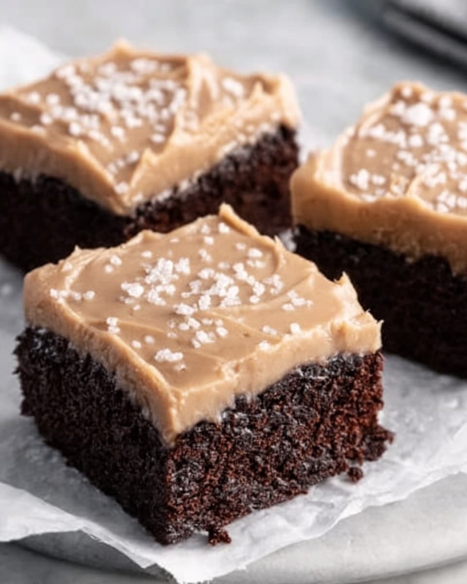 The image shows three square pieces of chocolate cake with a thick layer of light brown frosting on top, sprinkled with small white sugar crystals. The cake layers are dark and moist, while the frosting appears smooth and creamy. The pieces are arranged on white parchment paper, placed on a white marbled surface. photo taken with an iphone --ar 4:5 --v 7