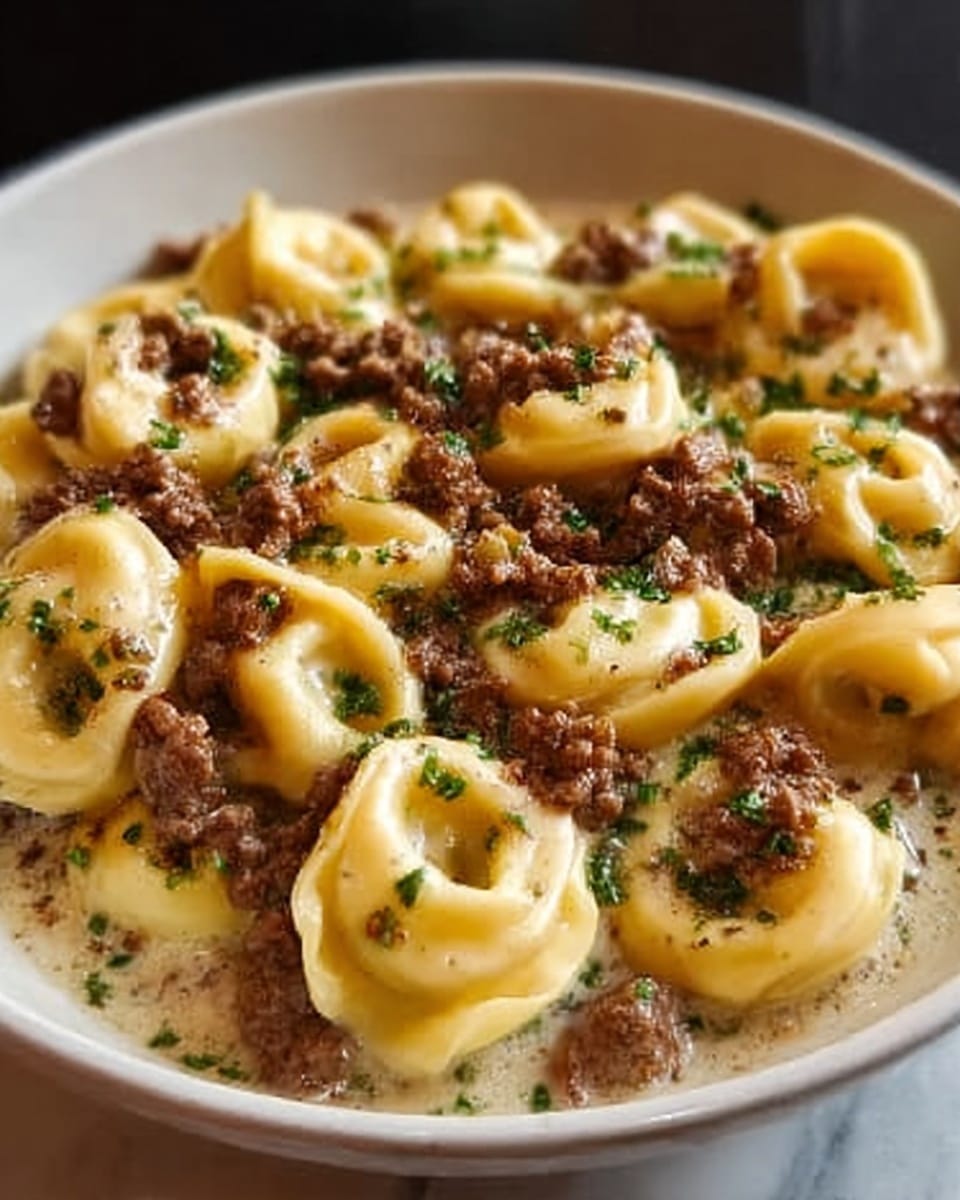 A white bowl filled with tortellini pasta covered in a creamy sauce, topped with ground beef pieces scattered evenly, and sprinkled with small green herb leaves. The pasta is golden yellow and folded in small ring shapes, sitting in the creamy light sauce, while the beef adds a rich brown color contrast. The surface beneath the bowl features a white marbled texture. Photo taken with an iphone --ar 4:5 --v 7