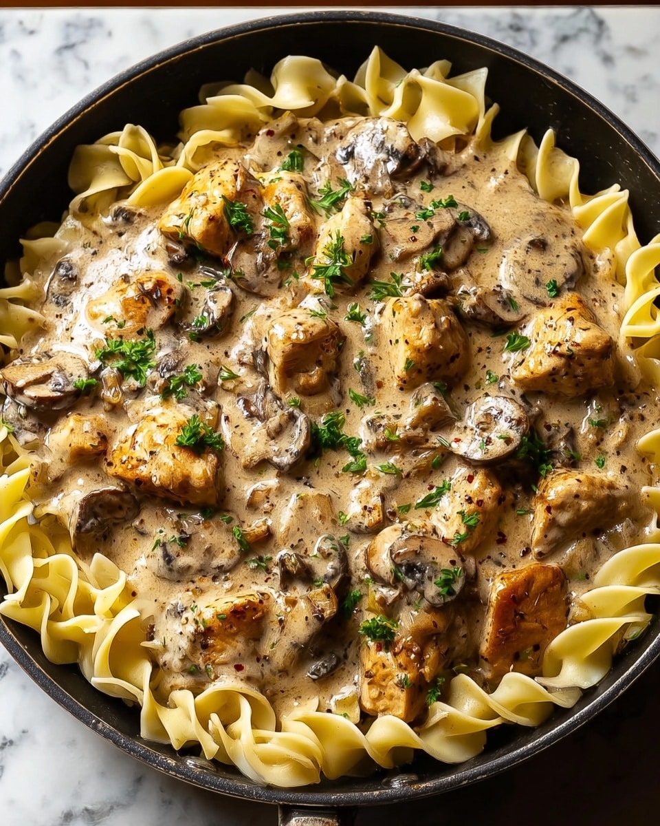 A skillet filled with wide, light cream-colored egg noodles as the base layer, topped with chunks of browned chicken pieces simmered in a thick, creamy mushroom sauce that has a beige color with dark specks from herbs and spices. Sliced mushrooms are scattered evenly throughout the sauce, adding a soft texture and darker brown color. The dish is garnished with small bits of fresh green parsley sprinkled on top, giving a fresh touch of color contrast against the creamy sauce. The skillet sits on a white marbled surface. Photo taken with an iphone --ar 4:5 --v 7