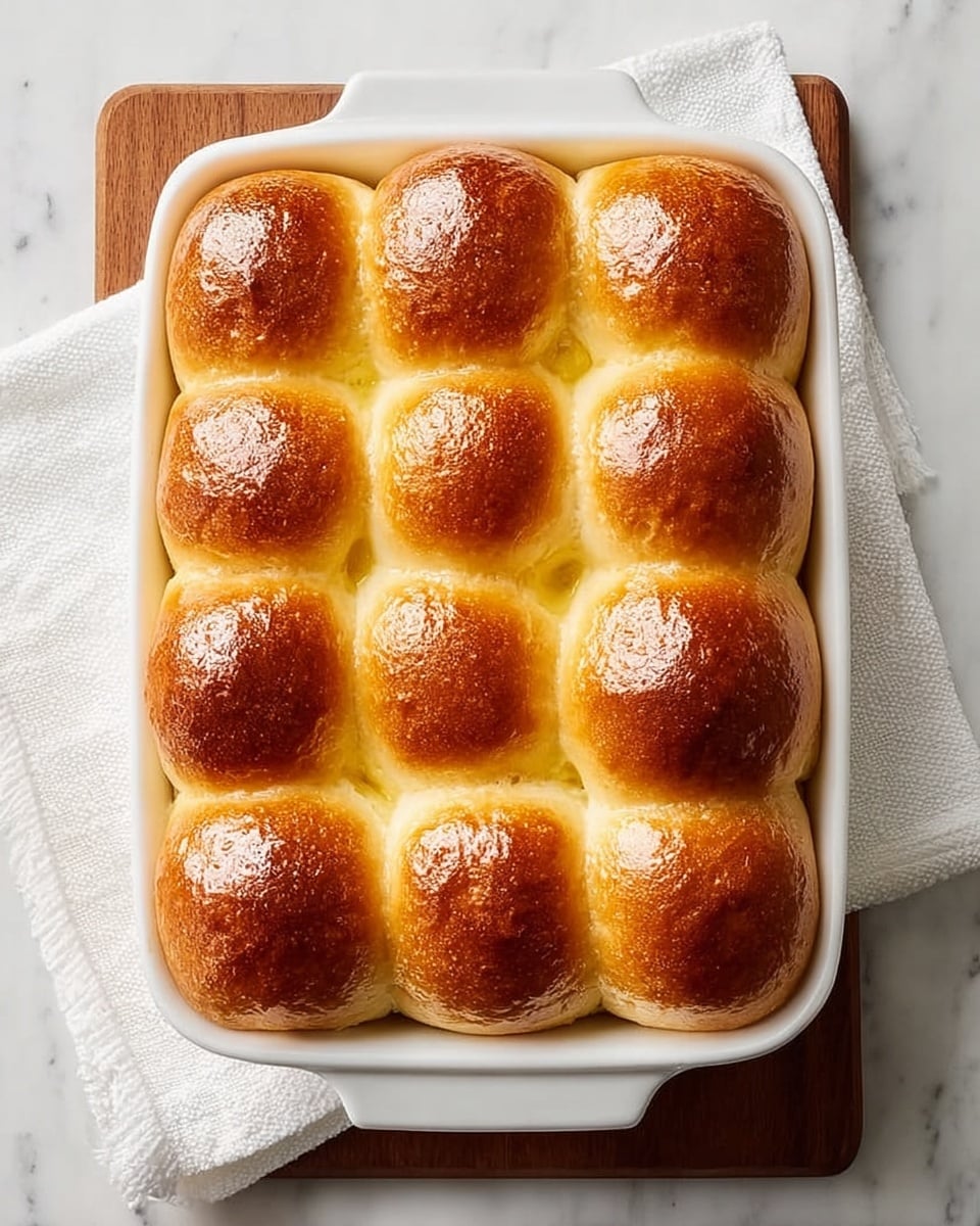 A white rectangular baking dish holds twelve soft bread rolls arranged in a 3 by 4 grid, each roll with a shiny golden brown top and a light yellow base. The rolls are touching each other, showing slightly rounded tops with a smooth texture and a glossy finish. The dish is placed on a wooden board, which sits on a white cloth over a white marbled surface. photo taken with an iphone --ar 4:5 --v 7