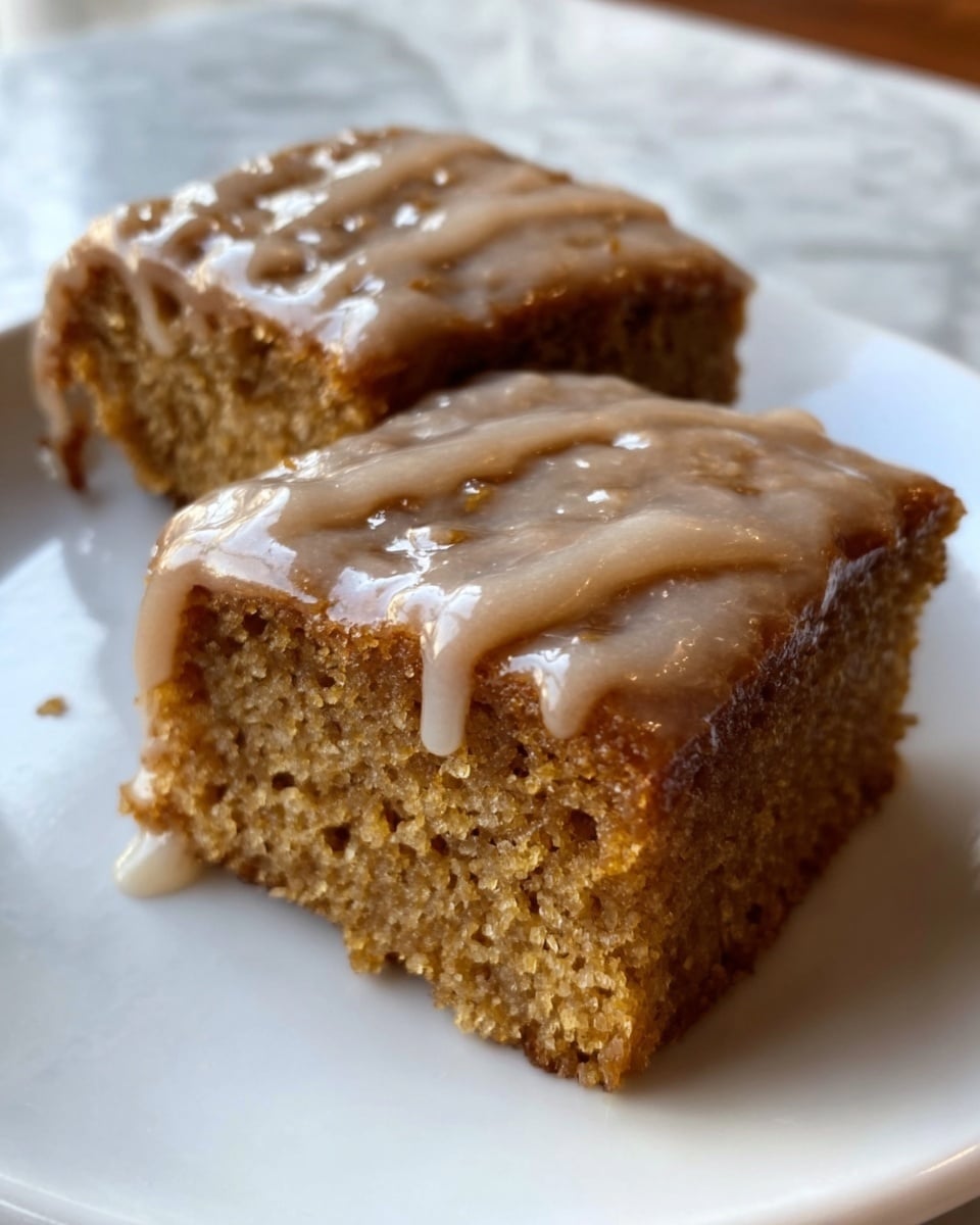 The image shows two square pieces of moist cake placed side by side on a white plate, each topped with a light brown glaze that looks smooth and shiny, slightly dripping down the sides. The cake layers are thick and dense, with a crumbly texture visible on the edges, and the color is a warm golden brown with small darker spots inside, indicating spices or nuts. The plate rests on a white marbled surface, with soft lighting highlighting the moistness and glaze on the cake. Photo taken with an iphone --ar 4:5 --v 7