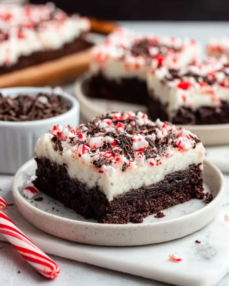 A close-up shot of a square piece of chocolate dessert with three layers: the bottom layer is thick and dark chocolate cake with a soft texture, the middle layer is creamy white frosting, and the top layer is decorated with crushed red and white candy canes and small pieces of dark chocolate shavings spread evenly. The dessert sits on a white round plate placed on a wooden surface. In the background, more dessert pieces are visible slightly out of focus, alongside a small white bowl filled with chocolate shavings and a candy cane lying on the wooden surface. Photo taken with an iphone --ar 4:5 --v 7