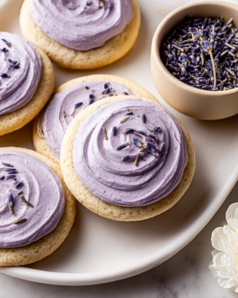 The image shows several round sugar cookies with one thick layer of smooth, light purple frosting swirled on top, decorated with small dried purple lavender flowers. The cookies are placed on a white oval plate, with more lavender flowers in a beige bowl on the side and a white flower on the white marbled surface. The lighting is gentle, highlighting the soft texture of the frosting and the crumbly look of the cookies. photo taken with an iphone --ar 4:5 --v 7