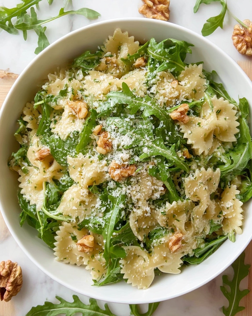 A white bowl filled with two main layers: a bottom layer of bowtie pasta that is pale yellow with a smooth texture and a top layer of fresh arugula leaves that are bright green and slightly wilted. Small bits of light brown walnuts are scattered evenly throughout, along with a light dusting of grated white cheese that adds a slight grainy texture on top. The bowl is placed on a white marbled surface, with some loose arugula leaves and walnuts around it. photo taken with an iphone --ar 4:5 --v 7