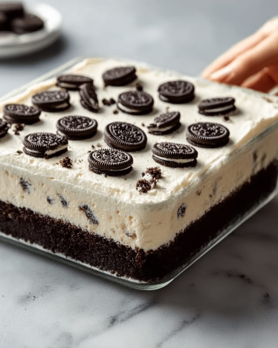 A square-shaped layered dessert in a clear glass dish, showing three distinct layers: the bottom is a thick, dark brown crumbly layer, the middle consists of a creamy light beige layer with small cookie pieces mixed in, and the top is a smooth white layer sprinkled with whole dark chocolate sandwich cookies evenly spaced. The dessert is on a white marbled surface, with a woman's hand reaching toward it from the side. photo taken with an iphone --ar 4:5 --v 7