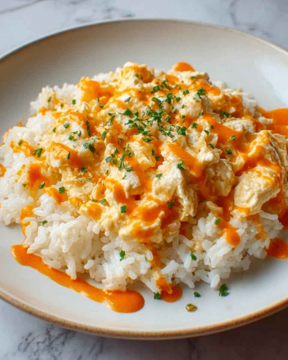 A round white plate holds a dish with two main layers. The bottom layer is a bed of plain white rice, soft and fluffy. On top, there is a thick layer of creamy scrambled eggs mixed with small bits of chicken or tofu, light yellow with a soft texture. The dish is drizzled with a bright orange sauce in thin lines across the eggs and rice. Small green parsley bits are sprinkled lightly over the top, adding color contrast. The plate rests on a white marbled surface. photo taken with an iphone --ar 4:5 --v 7