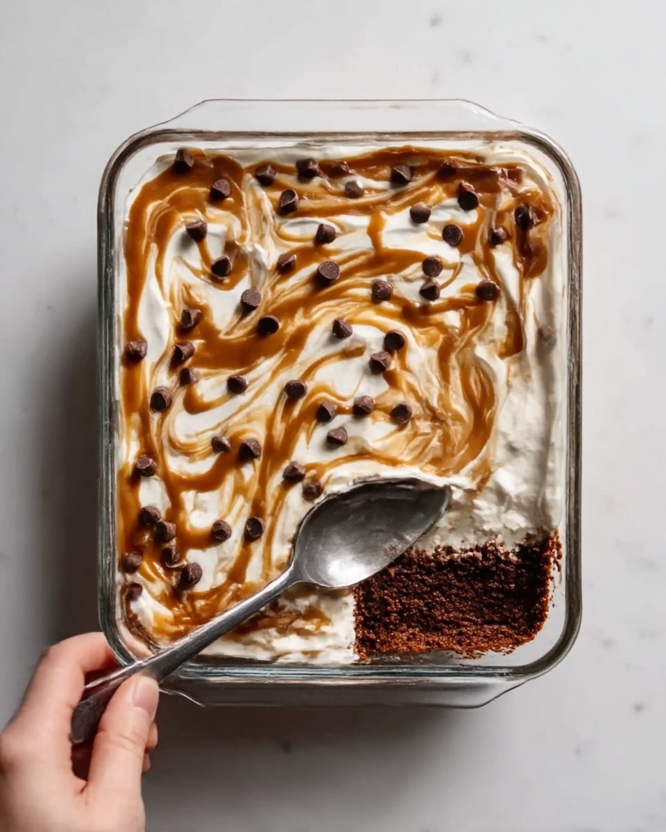 A square glass dish filled with a layered dessert topped with a white creamy layer spread evenly across the surface. On top of the white layer, there are irregular swirls of light brown caramel sauce creating a marbled effect and scattered dark chocolate chips evenly spread out. A spoon is inserted into the dessert on the right side, revealing the dark brown, dense bottom layer underneath the creamy top. The dish is placed on a white marbled surface, and a woman's hand is holding the spoon. Photo taken with an iphone --ar 4:5 --v 7