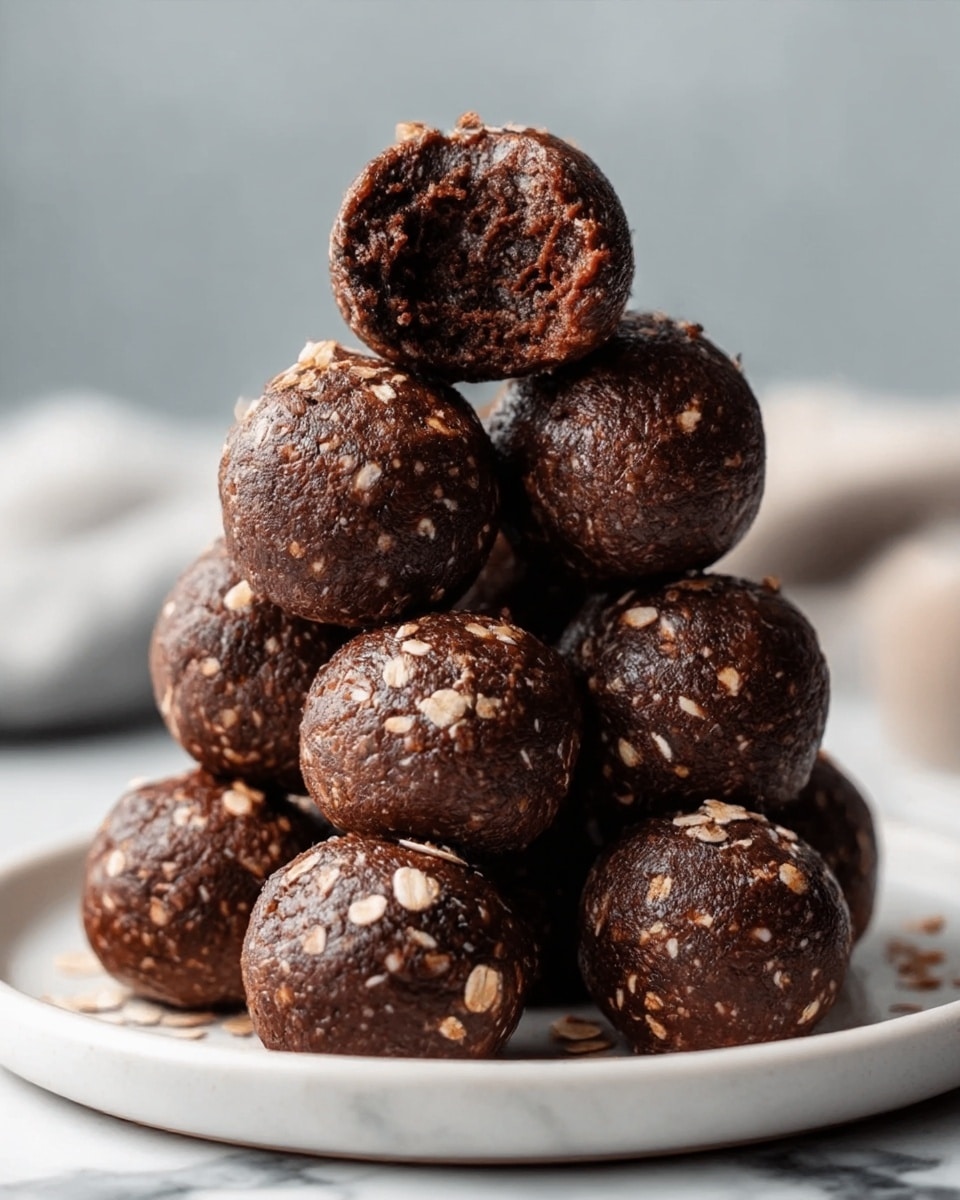 A white plate with a pile of eight round chocolate balls stacked in a pyramid shape. Each ball is dark brown, smooth, and glossy, with small pieces of oats sprinkled on top, giving a rough texture. The top ball has a bite taken out of it, showing a dense, moist, and slightly crumbly inside that is dark chocolate brown. The background is softly blurred with neutral tones, and the surface beneath the plate is a white marbled texture. Photo taken with an iphone --ar 4:5 --v 7