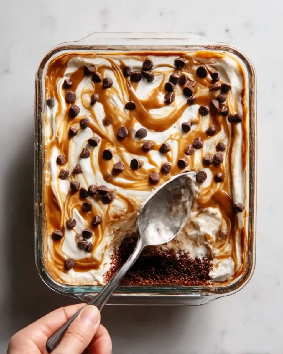 A glass container filled with a layered dessert. The bottom layer is dark brown, dense, and moist, likely chocolate cake or brownie. Above it is a thick, white layer of smooth cream or frosting. On top of the cream, there is a light brown swirl of caramel sauce, spread unevenly in circular patterns. Scattered over the caramel are many small, round, dark chocolate chips. A silver spoon with a clean handle is placed inside the container on the right side, partially buried in the dessert. The entire container is set on a white marbled surface. photo taken with an iphone --ar 4:5 --v 7