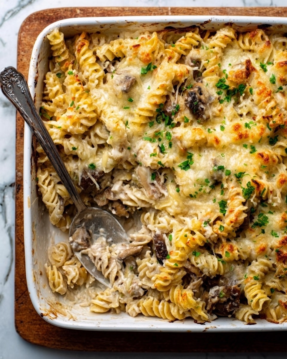 A close-up of a white rectangular casserole dish filled with baked pasta. The top layer is golden brown melted cheese with crispy edges and some green herb sprinkles. Underneath, there are spiraled pasta pieces mixed with chunks of browned meat and creamy sauce, visible where some pasta has been scooped out with a silver spoon resting on the right side of the dish. The dish sits on a wooden surface with a white marbled texture in the background, alongside some green herbs peeking in the corner. Photo taken with an iphone --ar 4:5 --v 7