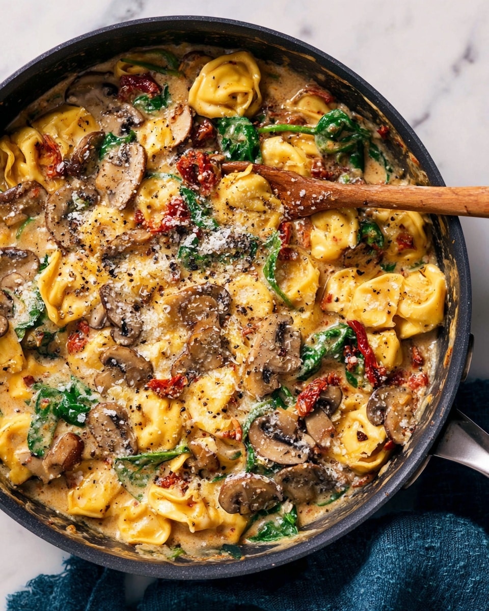 The image shows a close-up of a black pan filled with creamy tortellini pasta that has a rich yellow color and soft, pillowy texture. The dish is layered with sautéed brown mushrooms scattered throughout, wilted green spinach leaves adding freshness, and sun-dried tomatoes giving a deep red contrast. The sauce is thick, cheesy, and glossy, coating every piece of pasta and vegetable, with a sprinkling of grated cheese on top. A wooden spoon is stirring the contents, partially lifting some tortellini. The pan sits on a white marbled surface with a blue cloth napkin beside it. Photo taken with an iphone --ar 4:5 --v 7